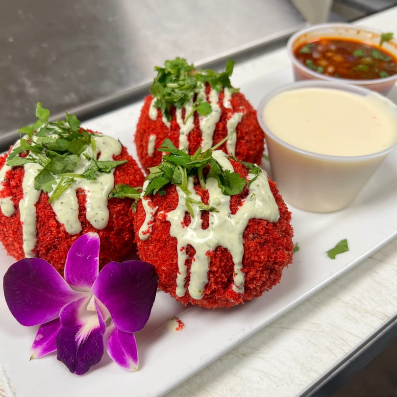 Three red fried balls topped with green sauce and cilantro, served with two dipping sauces and a purple orchid.