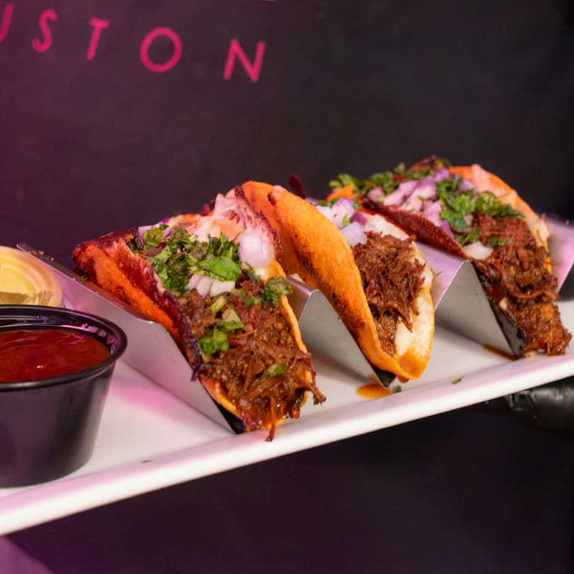 Three birria tacos with onions and cilantro on a white plate with red salsa.