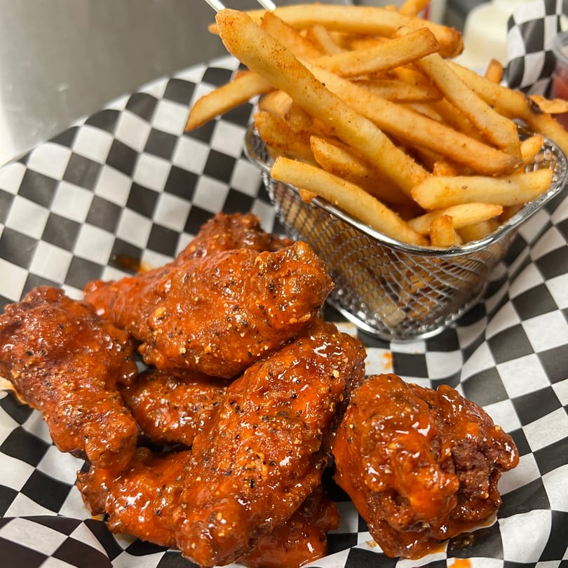 Red sauced chicken wings and crispy golden fries in a basket on checkered paper.