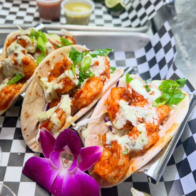 Three shrimp tacos with white sauce, green onions, and a purple orchid on a checkered tray.