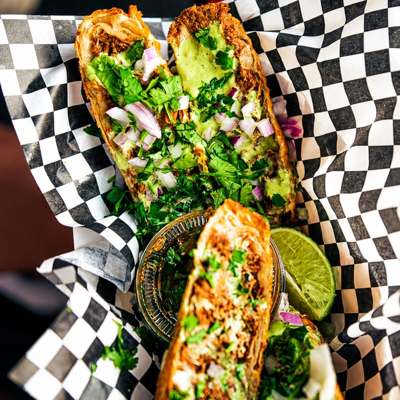 Two crispy tacos filled with meat, green sauce, cilantro, and red onions, served in checkered paper.